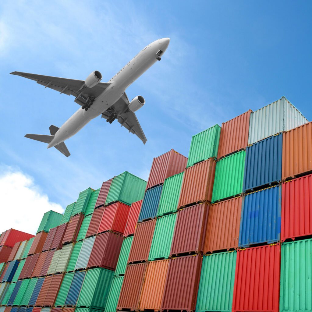 A large white cargo plane flying over a wall of stacked colorful shipping containers against a blue sky, representing global Air Freight logistics.
