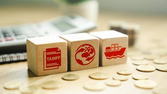 Three wooden blocks with red icons of a tariff document, a globe, and a cargo ship, placed near coins and a calculator.