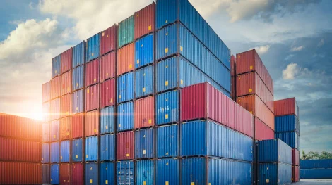 Towering stacks of red and blue shipping containers illuminated by an intense sunburst, illustrating the global trade context of HS Code vs UK Commodity Code customs comparisons.